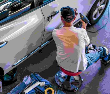 Man working on a New Car door in a garage, kneeling beside a white vehicle and using tools to inspect or repair it.