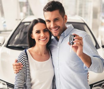 Smiling couple standing in front of a car at a dealership holding car keys, illustrating tips to Avoid Bad Credits and make smarter financial decisions when buying a vehicle.