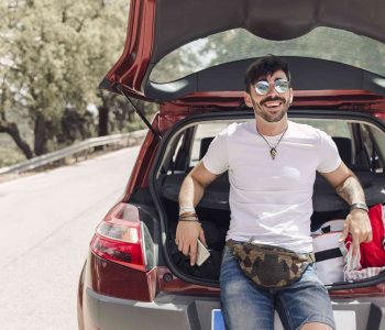Person sitting in the open trunk of a red Small Car parked on a sunny road with trees in the background.